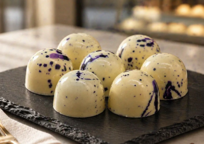 seven blueberry and glossy, dome-shaped white chocolates splattered and marbled with dark purple speckles, arranged on a black slate tray