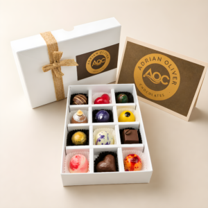 An open white gift box showcasing 12 colorful, hand-decorated artisan chocolates alongside a closed box tied with twine and an Adrian Oliver Chocolates branded card on a light background.
