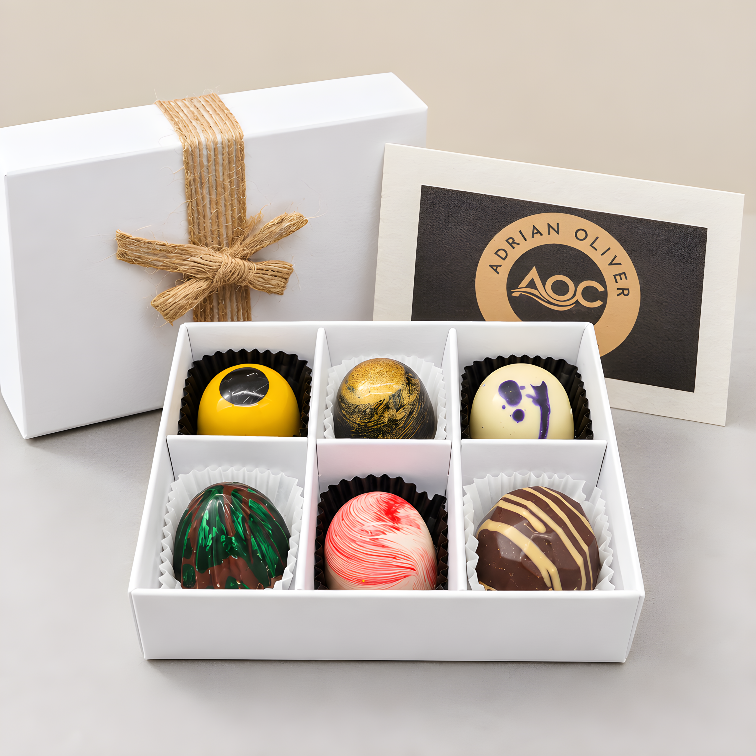 An open white gift box containing six handcrafted, colorful artisan chocolates, placed alongside a tied gift box lid and an Adrian Oliver Chocolates brand card.