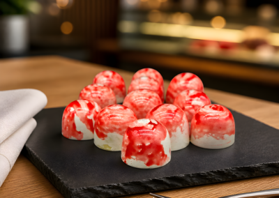 A collection of glossy, dome-shaped white chocolates featuring a red marbled pattern across their tops, arranged on a black slate tray set against a warm, softly lit background.