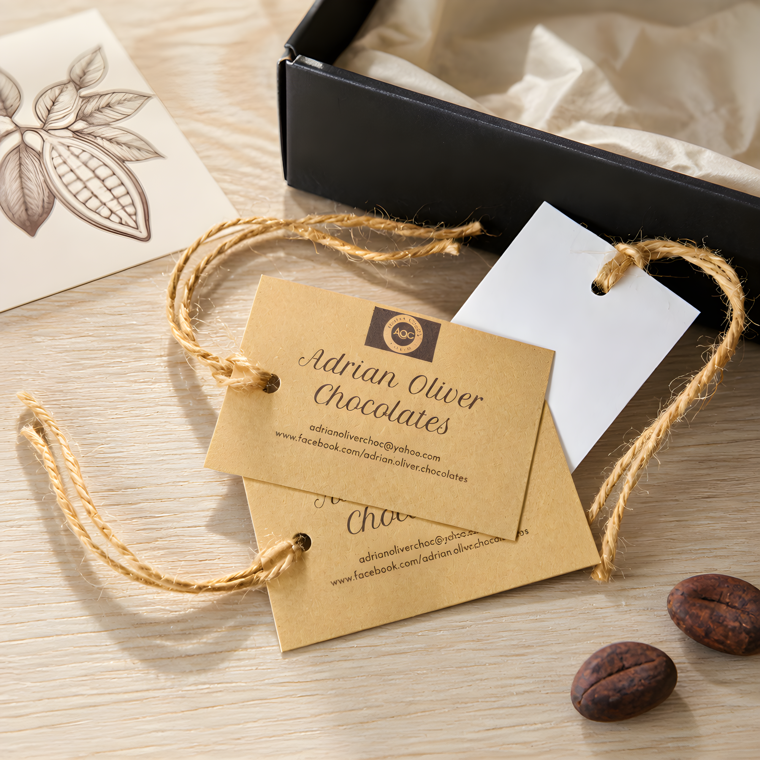 Two craft paper gift tags tied with twine strings showing contact details for "Adrian Oliver Chocolates" placed on a light wood surface. A white card with a hole punch, an open black gift box lined with tissue paper, an botanical cocoa pod illustration, and two cocoa beans rest nearby.